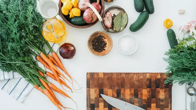 A stainless dog bowl surrounded by fresh vegetables, fish, and a small pile of supplement powder